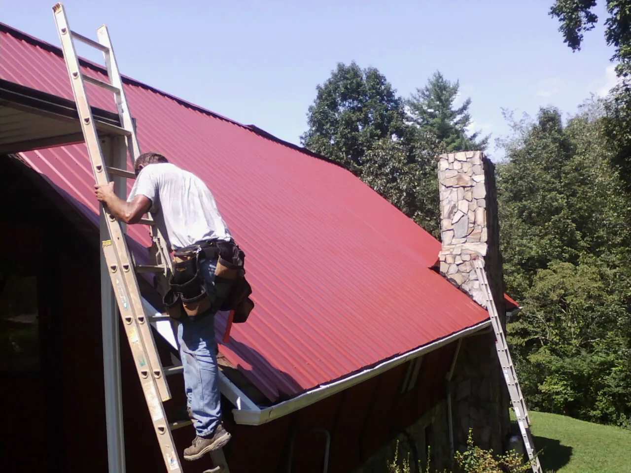 Roofer on a ladder beside a red metal roof and stone chimney — Soffit & Fascia Repair by Roofing Brothers in Folkston